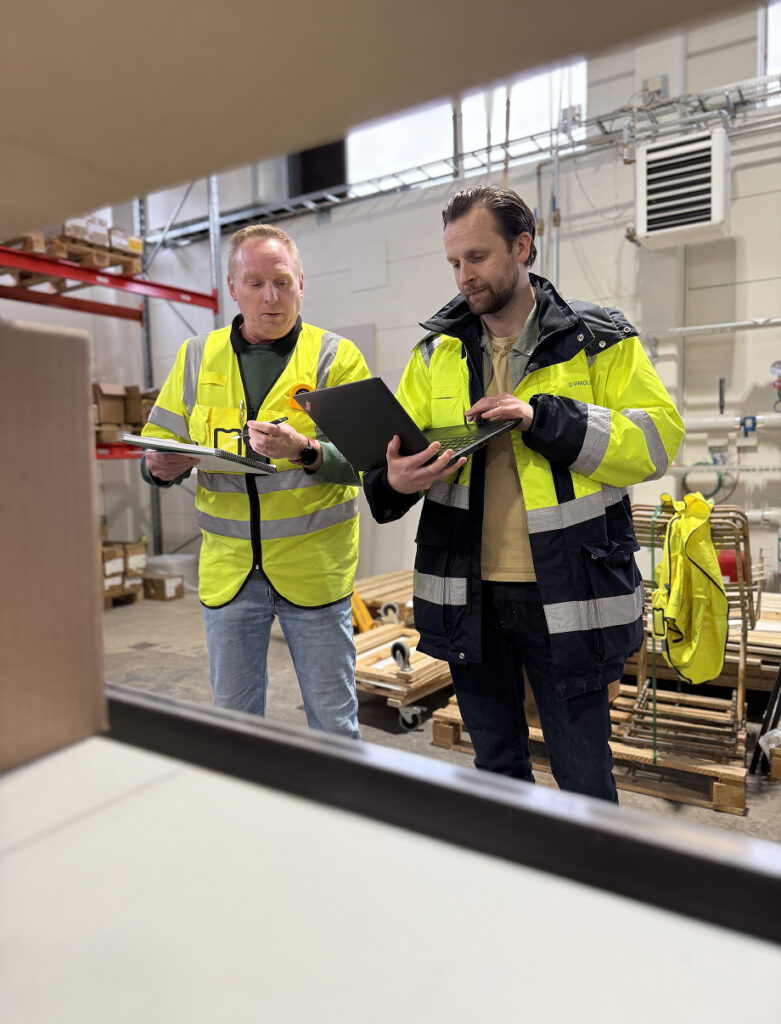 Two men in high-visibility jackets review documents and a laptop in a warehouse setting.